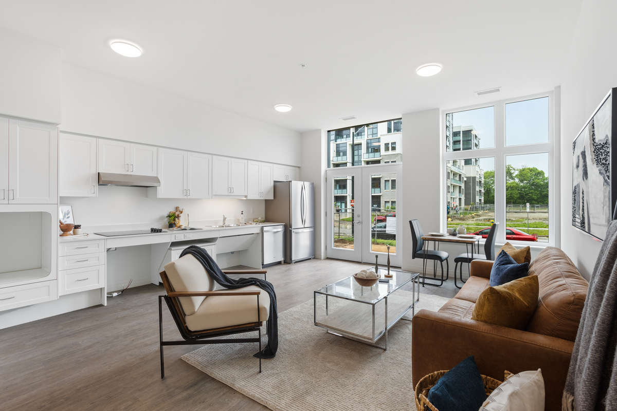Bright spacious modern living room with open concept kitchen, couch, coffee table, armchair and a dining table by a large window. Photo of a one-bedroom model suite apartment at Zerin Place on South Street at 370 South St. in London Ontario. A housing project by Zerin Development in VillageSoHo.